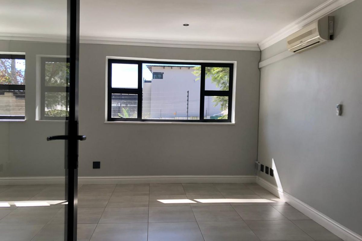 A bright, empty office space featuring tiled flooring, recessed downlights, a wall-mounted air conditioning unit, and modern black-framed windows.