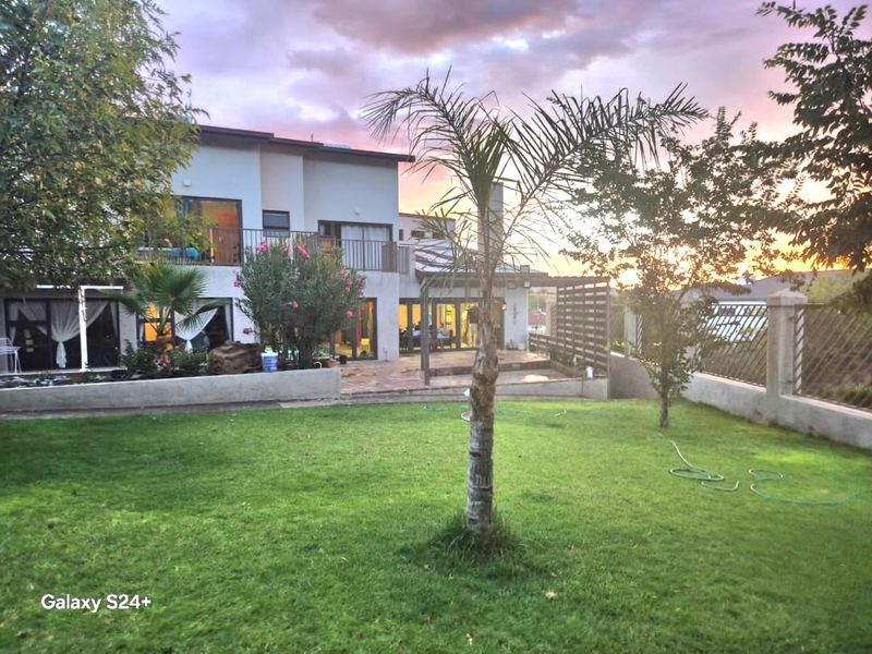 A rear view of the property featuring a landscaped lawn, a paved patio area with a pergola, and an upper-level balcony at sunset.