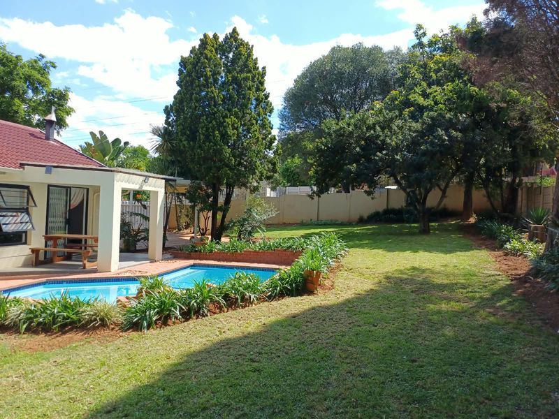 Spacious backyard featuring a swimming pool with brick coping, a covered patio area, and a well-maintained lawn surrounded by mature trees and perimeter walls.
