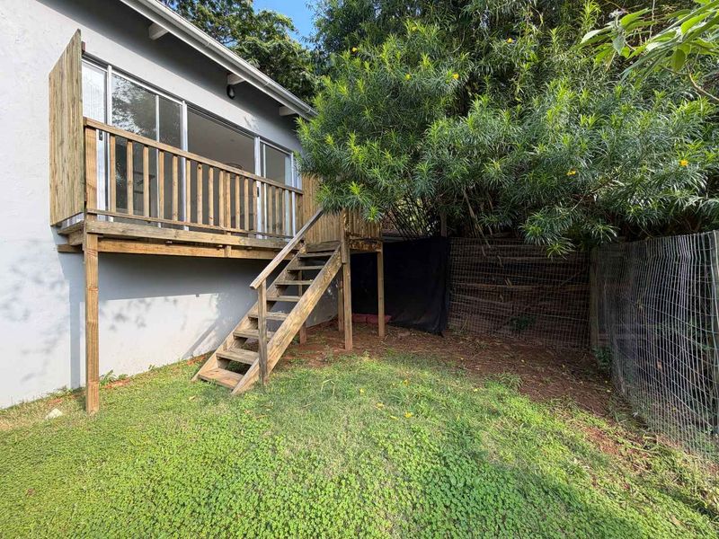 Rear view of the cottage featuring a raised wooden deck with staircase access, sliding glass doors, and a private garden area with perimeter fencing.