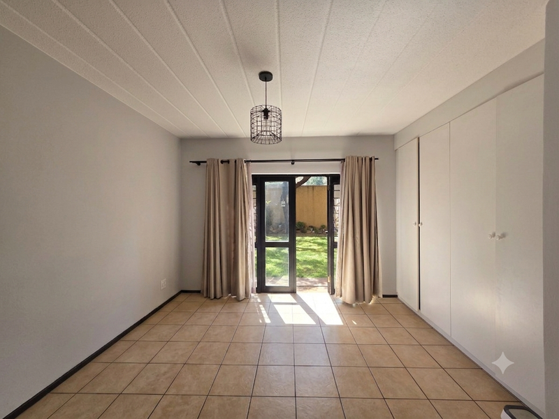 A bright, empty bedroom featuring neutral tiled flooring, ample built-in cupboard storage, and direct access to a private garden area through a glass door.