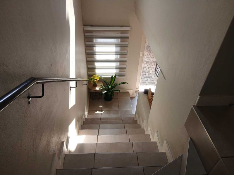 A bright, tiled staircase featuring a modern stainless steel handrail and a window with zebra blinds, leading down to the entrance area.