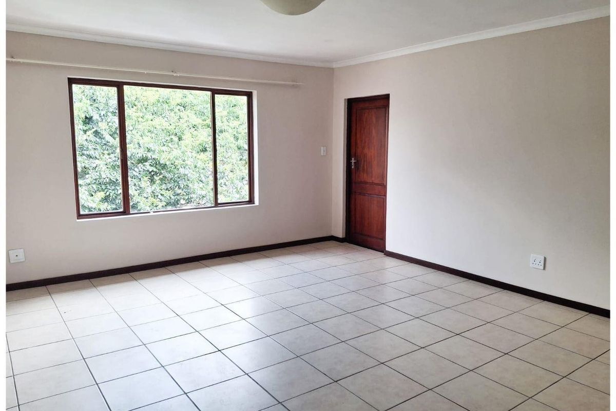 A bright, empty living room featuring neutral tiled flooring, a large window with garden views, and a solid timber door.