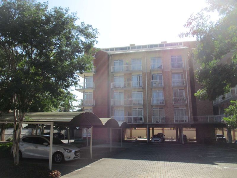 Exterior view of a multi-story apartment building featuring brick facade, private balconies, and covered parking bays on a paved driveway.