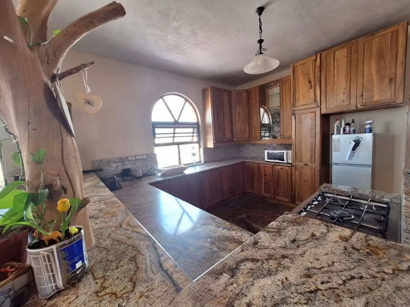 Spacious kitchen featuring warm wooden cabinetry, extensive granite countertops, a built-in gas hob, and a distinctive arched window with stone tile flooring.