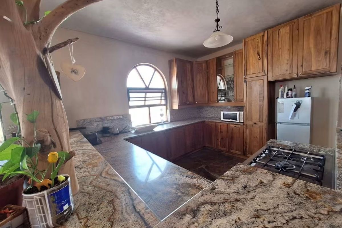 Spacious kitchen featuring warm wooden cabinetry, extensive granite countertops, a built-in gas hob, and a distinctive arched window with stone tile flooring.