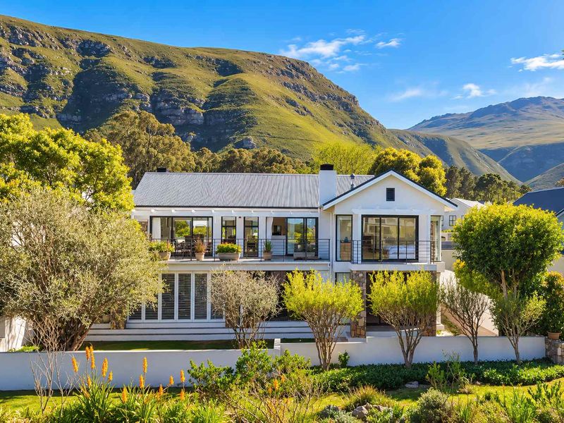 A beautifully maintained two-story residence featuring a corrugated metal roof, stone accents, and a spacious balcony overlooking a lush garden and mountain backdrop.