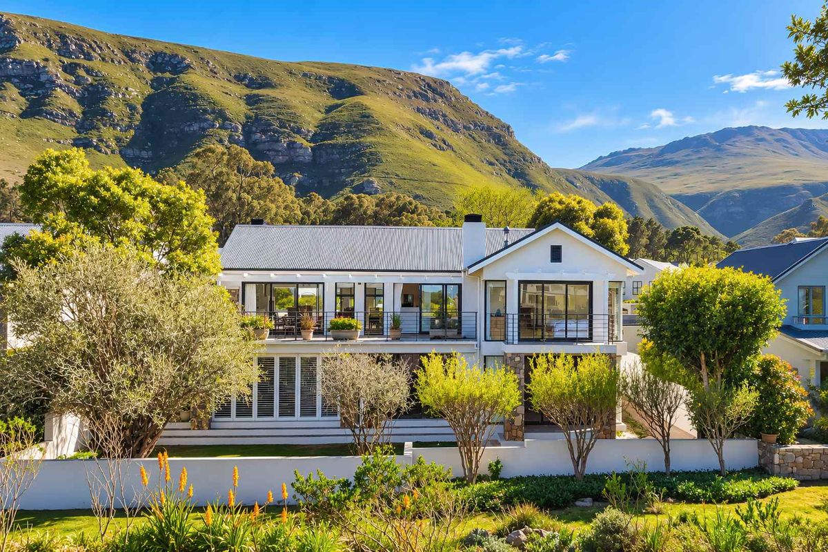A beautifully maintained two-story residence featuring a corrugated metal roof, stone accents, and a spacious balcony overlooking a lush garden and mountain backdrop.