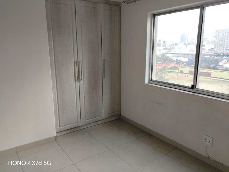 A bright, empty bedroom featuring neutral tiled flooring, a built-in wardrobe, and a window offering an urban view.