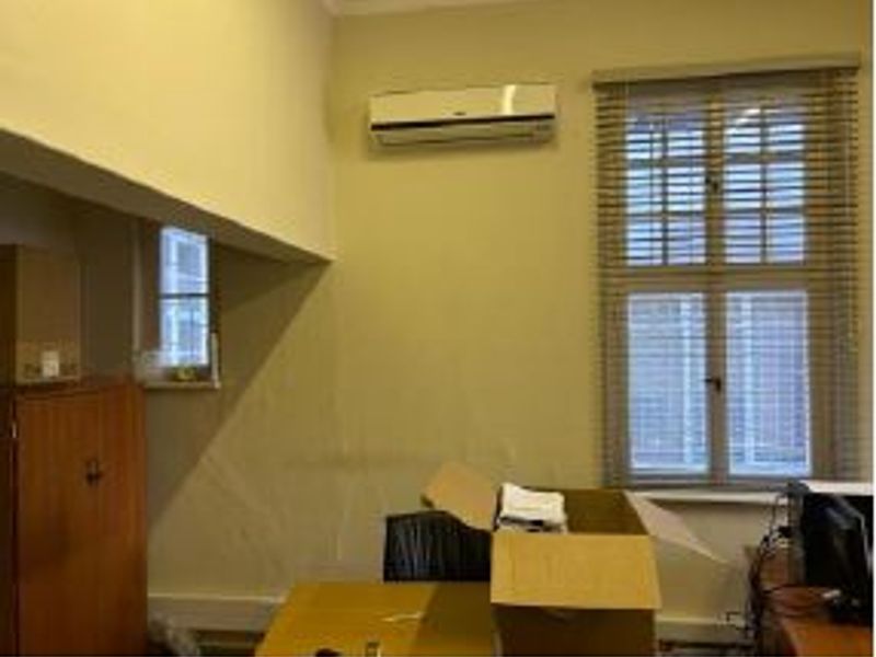 A functional office space featuring a wall-mounted air conditioning unit and large windows with fitted venetian blinds.