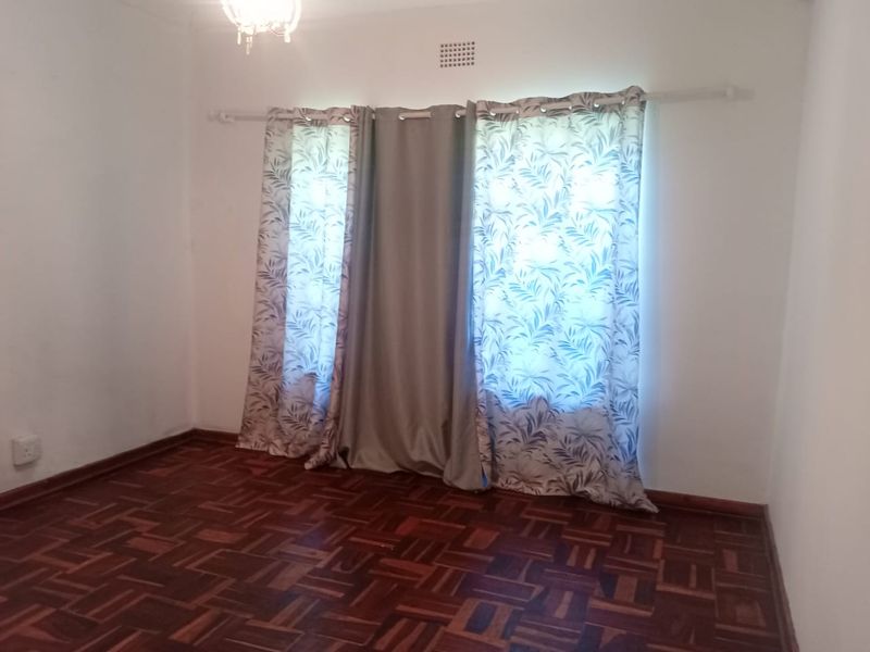 A bright, empty bedroom featuring classic parquet wood flooring, neutral walls, and a window fitted with a curtain rod.