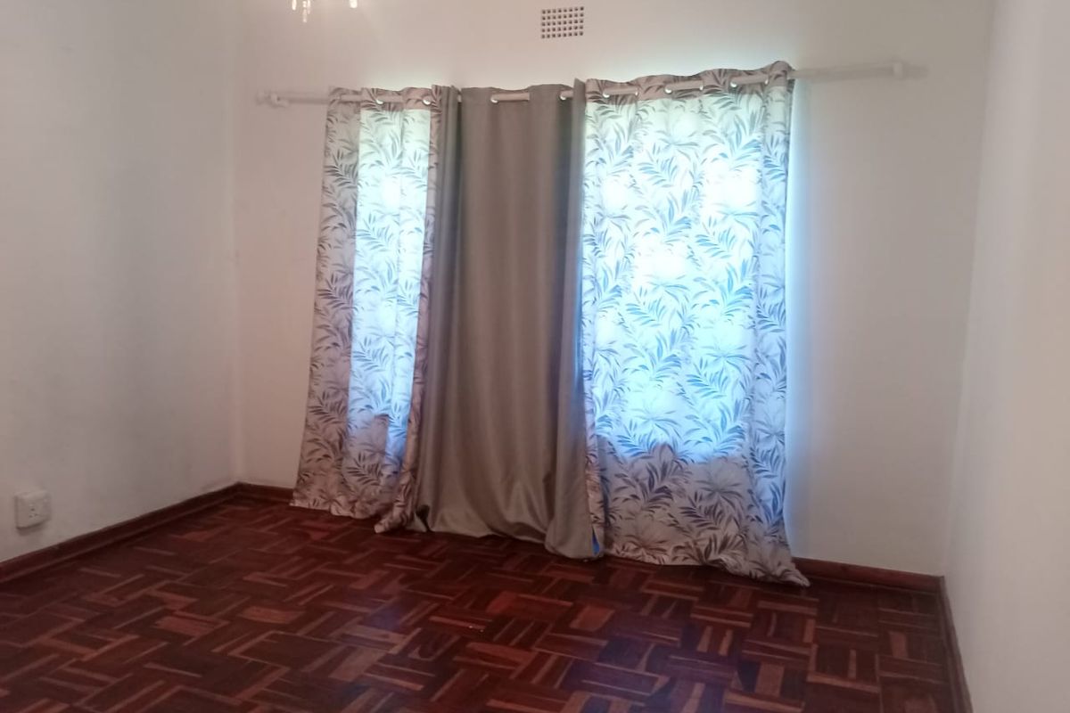 A bright, empty bedroom featuring classic parquet wood flooring, neutral walls, and a window fitted with a curtain rod.