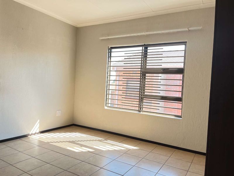 A bright, empty bedroom featuring neutral tiled flooring, textured walls, and a window fitted with security bars.