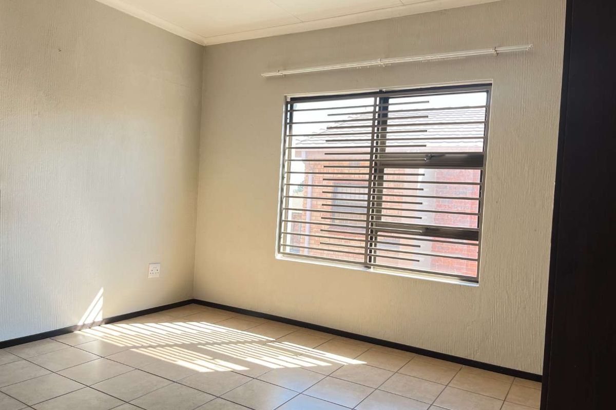 A bright, empty bedroom featuring neutral tiled flooring, textured walls, and a window fitted with security bars.