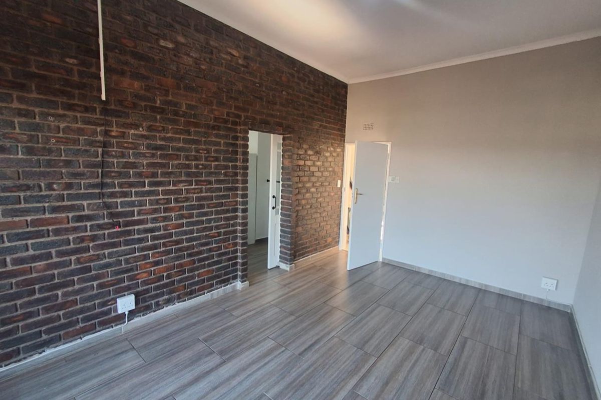 A bright, empty room featuring a distinctive exposed brick feature wall, modern wood-look tiled flooring, and neutral painted walls.
