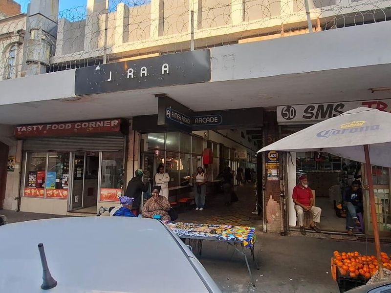 Street-level view of a commercial arcade entrance featuring multiple storefronts, a concrete awning, and security perimeter fencing.