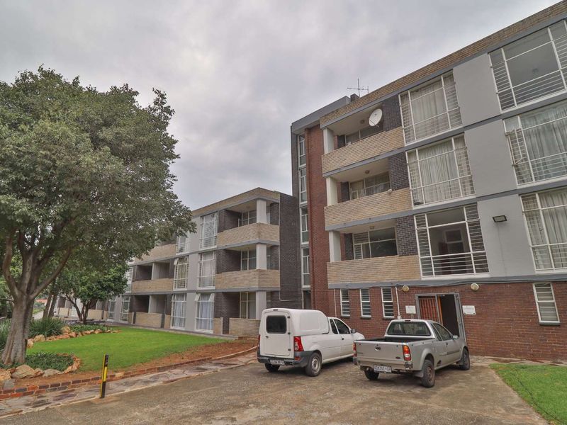 Exterior view of a multi-story apartment complex featuring brick facade, private balconies, and a paved driveway with landscaped garden area.