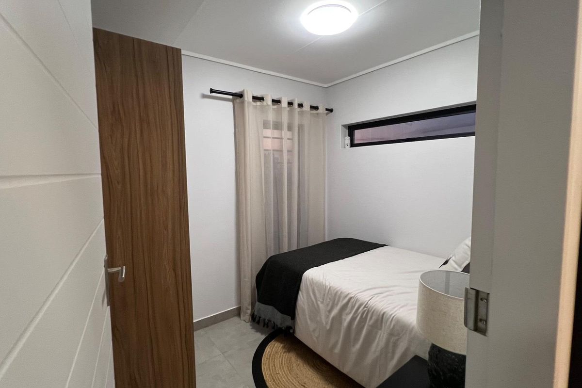 A compact, modern bedroom featuring neutral tones, tiled flooring, a high-set horizontal window, and a wood-finish door.