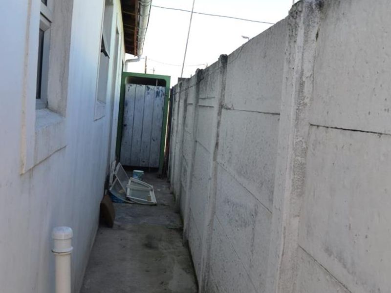 A narrow side passage featuring a concrete boundary wall, drainage downpipe, and a weathered rear access door.