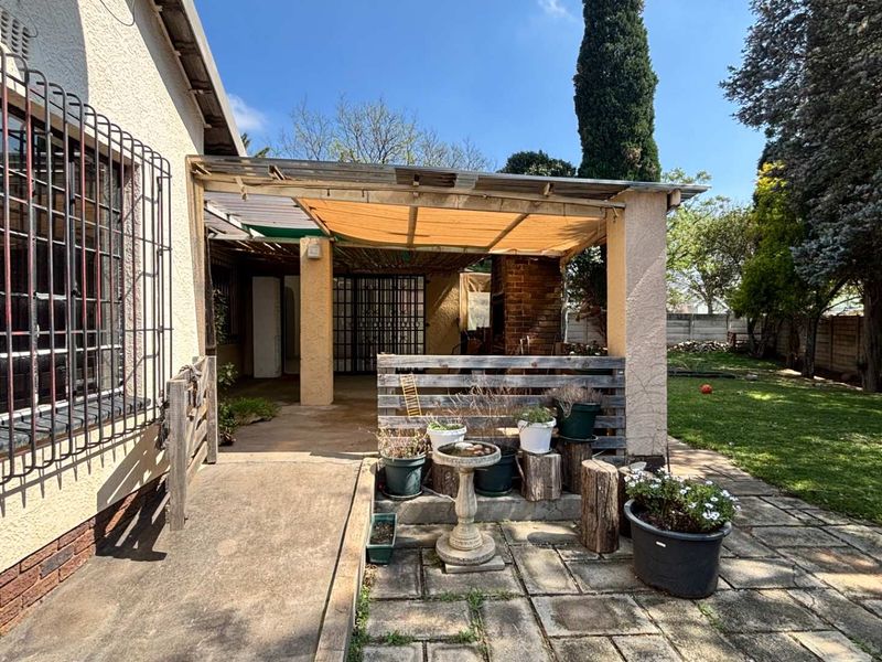A covered outdoor patio area featuring a brick fireplace, paved flooring, and security bars, overlooking a private garden space.