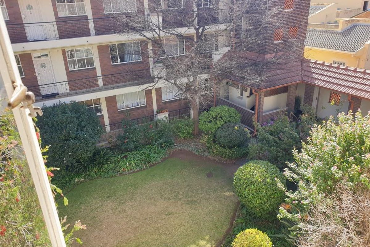 An elevated view overlooking a communal garden area with established landscaping, surrounded by a multi-story brick apartment building with balconies.