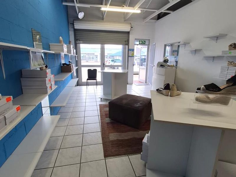 An open-plan office or retail space featuring exposed steel roof trusses, ceramic tiled flooring, and built-in wall shelving with a roller shutter entrance.