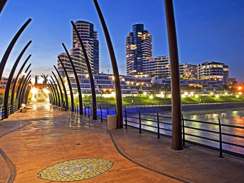 A scenic evening view of the coastal promenade featuring architectural pillars and decorative paving, overlooking the ocean and nearby apartment buildings.