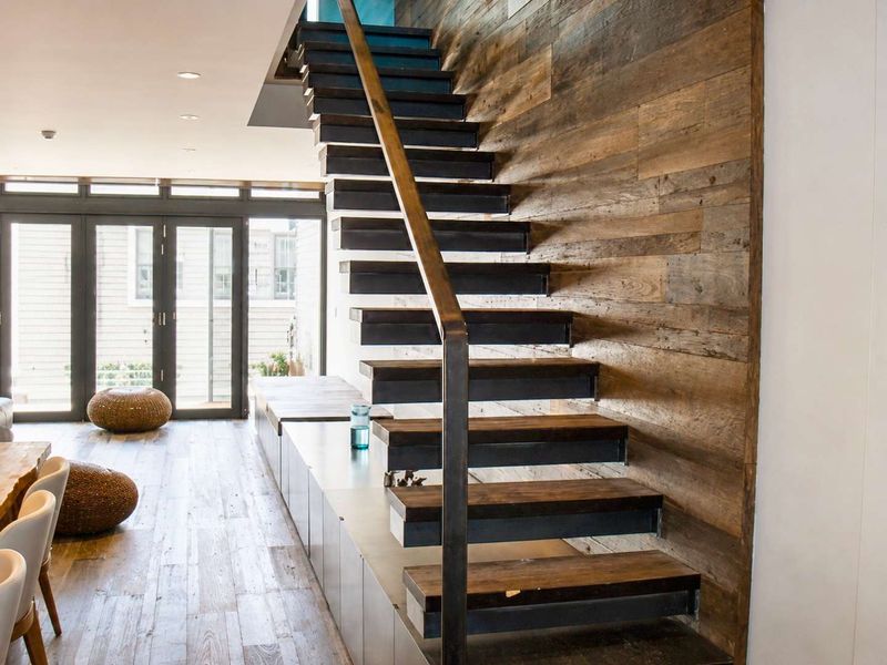 Modern open-plan living area featuring a striking floating staircase, reclaimed timber wall cladding, custom built-in cabinetry, and seamless access to the outdoor space.