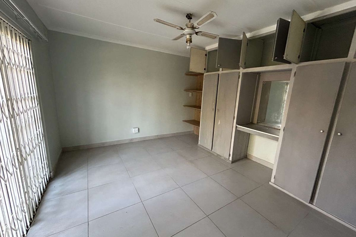 A spacious bedroom featuring neutral tiled flooring, built-in cupboards with a vanity area, and a ceiling fan, offering a blank canvas for personalization.
