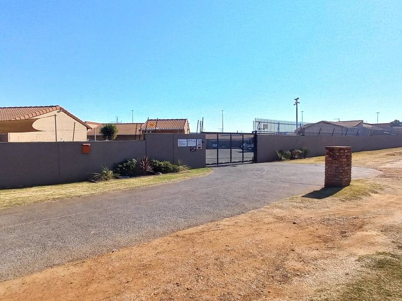 Front exterior view of the townhouse complex entrance, featuring a perimeter wall, security gate, and paved driveway access.