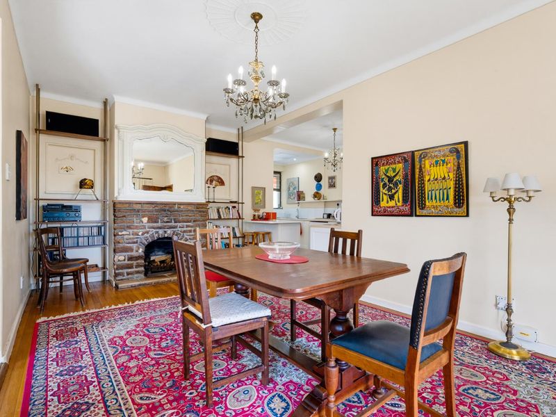 Open plan dining area featuring a classic brick fireplace, ornate ceiling rose, chandelier lighting, and polished hardwood flooring, with a view into the kitchen.