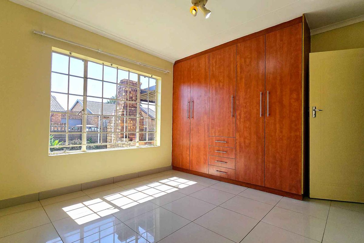 A bright bedroom featuring large ceramic floor tiles, a built-in wooden wardrobe, and a large window providing natural light.