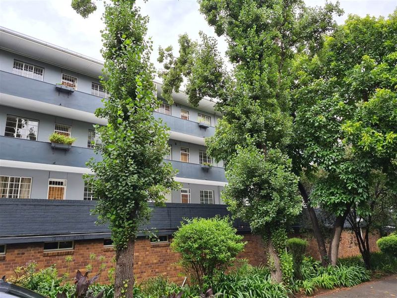 Exterior view of a multi-story apartment building featuring a brick boundary wall, mature trees, and private balconies.