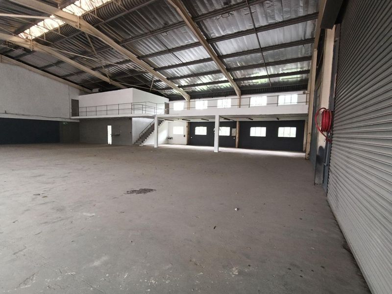 Spacious industrial warehouse featuring a mezzanine level, high exposed steel roof trusses, concrete flooring, and a large roller shutter door for easy access.