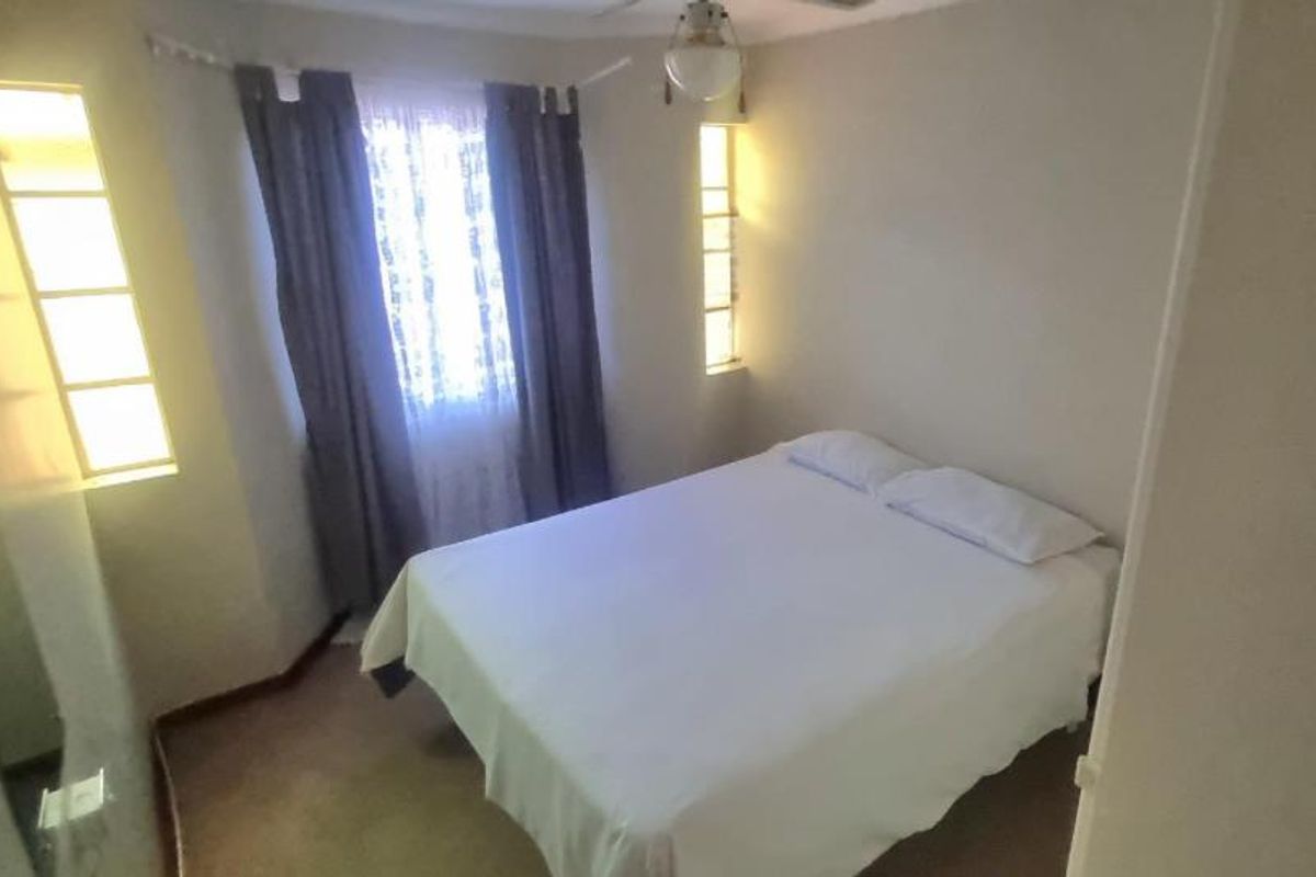 A compact bedroom featuring neutral walls, carpeted flooring, and natural light provided by two windows with curtains.