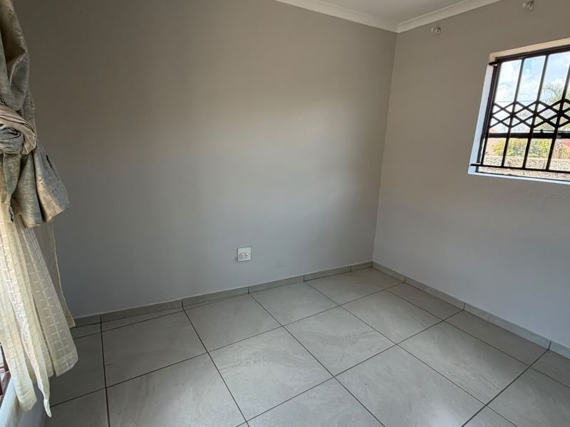A clean, empty bedroom featuring neutral tiled flooring, light-colored walls, and windows fitted with security bars.