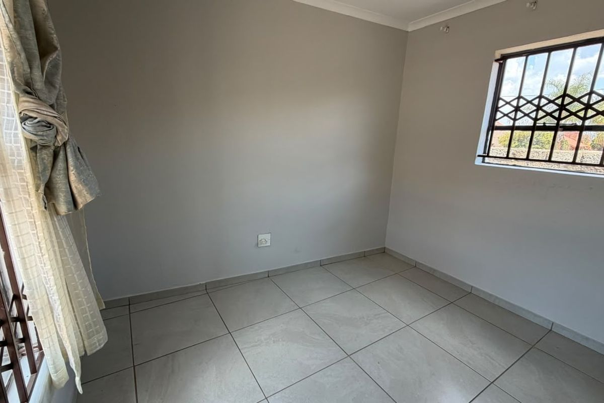 A clean, empty bedroom featuring neutral tiled flooring, light-colored walls, and windows fitted with security bars.