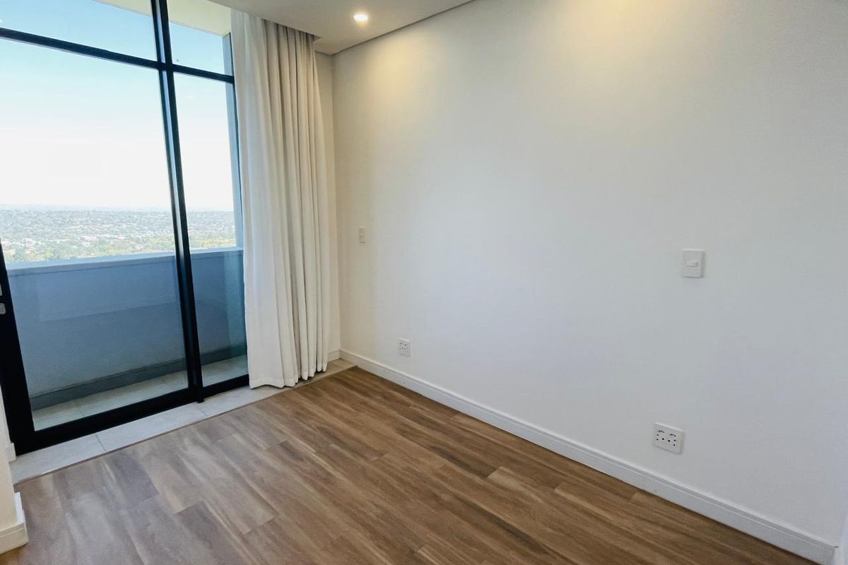 A bright, empty bedroom featuring modern laminate flooring, recessed lighting, and a large glass sliding door providing access to a balcony with expansive views.