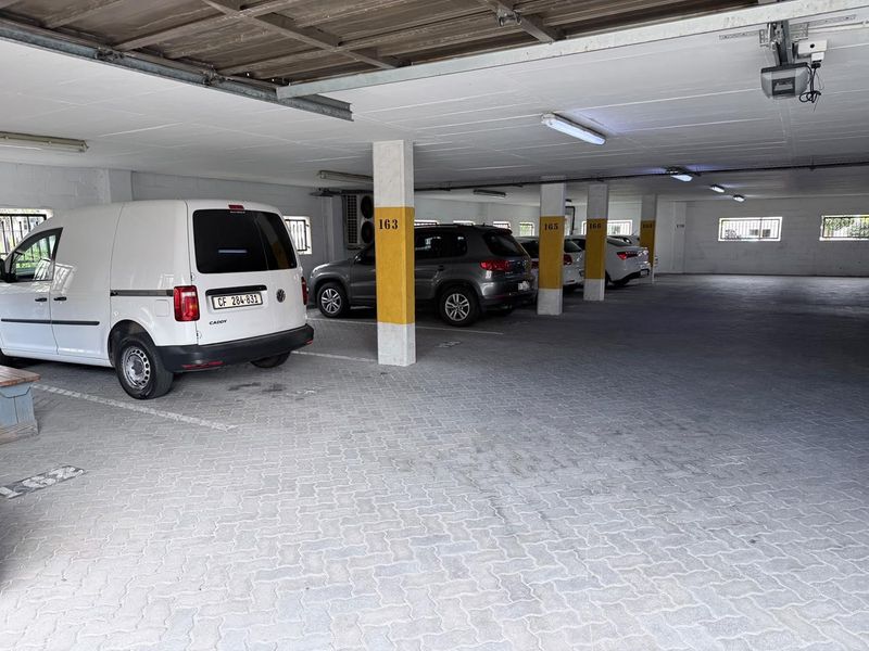 Covered parking garage area featuring paved flooring, structural support columns, and overhead fluorescent lighting.