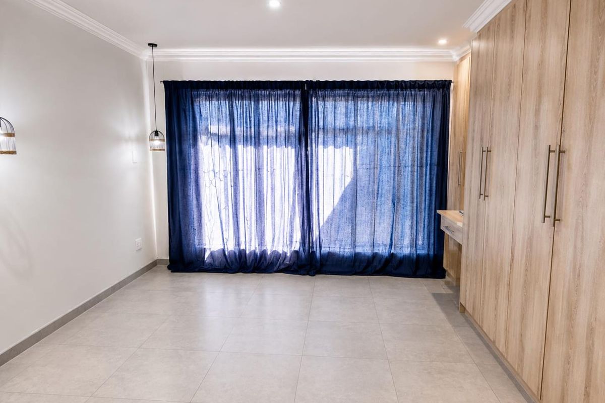 A bright, modern bedroom featuring neutral tiled flooring, extensive built-in wooden cabinetry with a vanity section, and elegant crown molding.