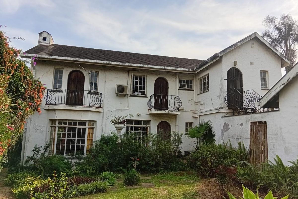 Rear elevation of a two-story house featuring multiple wrought iron balconies, arched doorways, and a tiled roof, set within a mature garden.