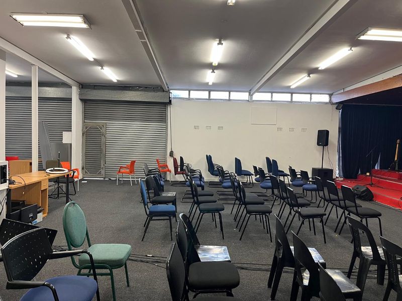 A large open-plan multi-purpose hall featuring a raised stage area, industrial roller shutter doors, and overhead fluorescent lighting.