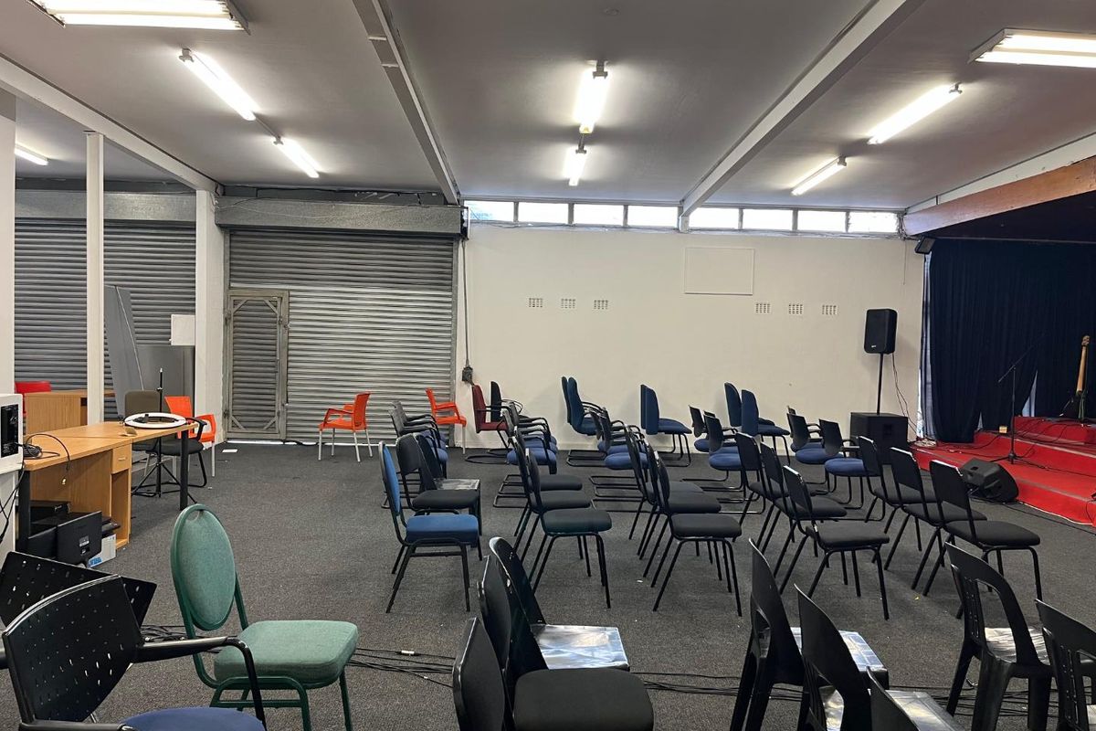A large open-plan multi-purpose hall featuring a raised stage area, industrial roller shutter doors, and overhead fluorescent lighting.