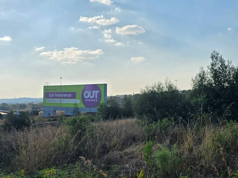 A view of a vacant residential plot featuring natural vegetation and perimeter fencing, with a large billboard visible in the background.