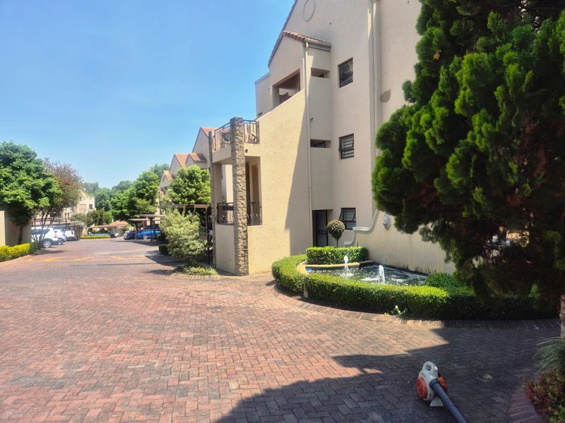 Exterior view of the apartment complex featuring a brick paved driveway, landscaped garden with a water fountain, and building facade with balconies.