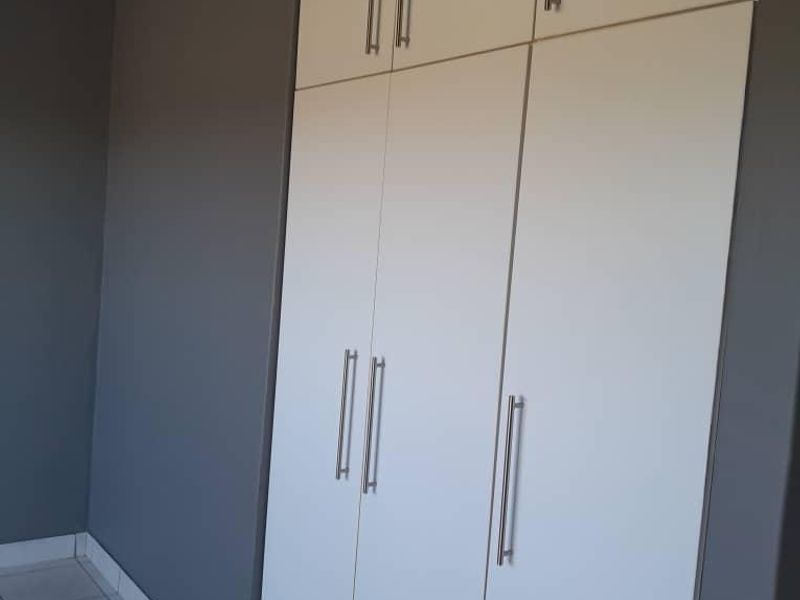 A clean, empty bedroom featuring modern grey walls, white built-in cupboards with silver handles, and neutral tiled flooring.