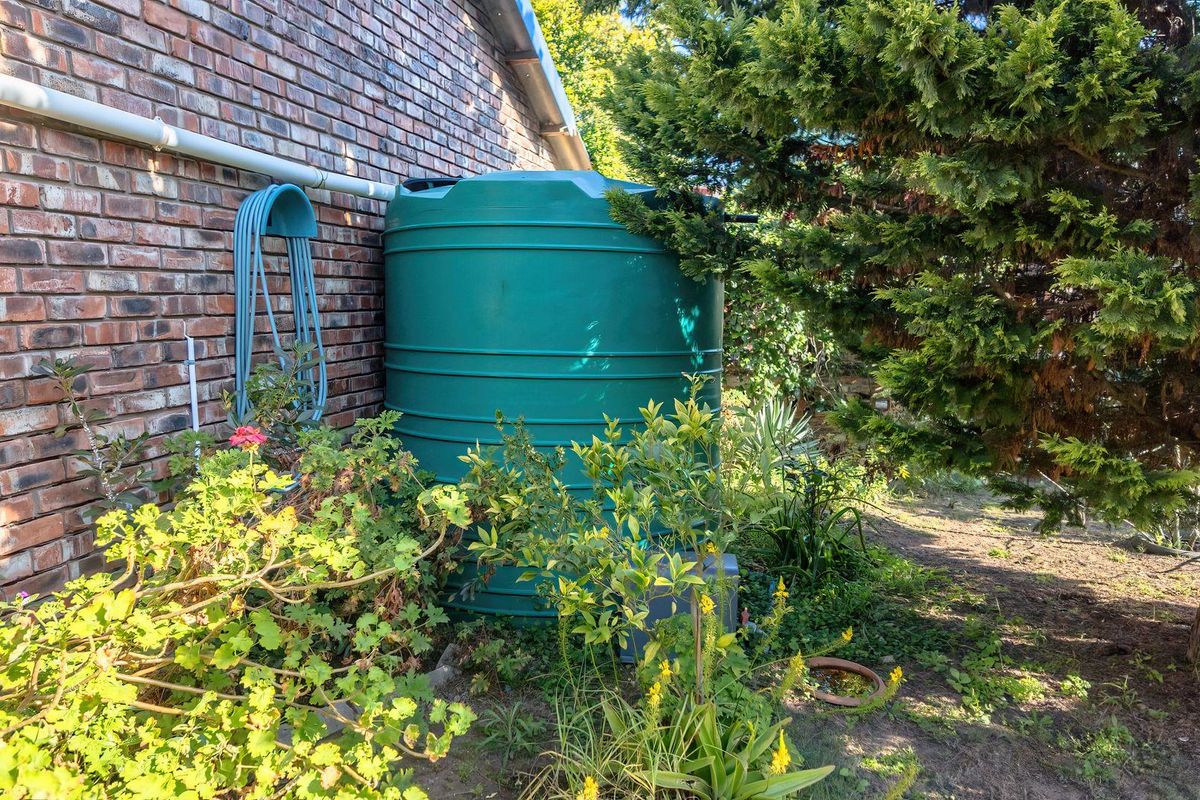 A side view of the property featuring a large green water storage tank positioned against a brick exterior wall with established garden landscaping.