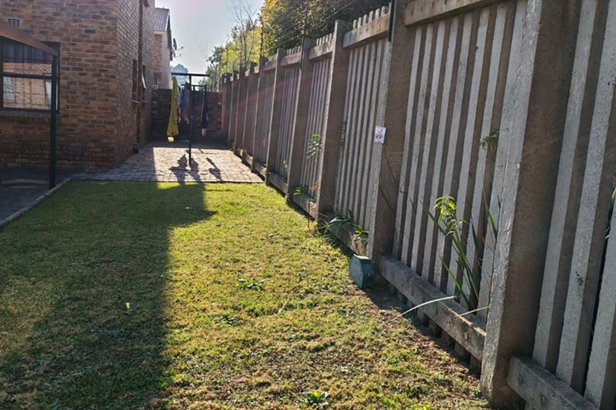 A rear garden area featuring a lawn, brick-paved pathway, and concrete palisade fencing topped with electric security wiring.