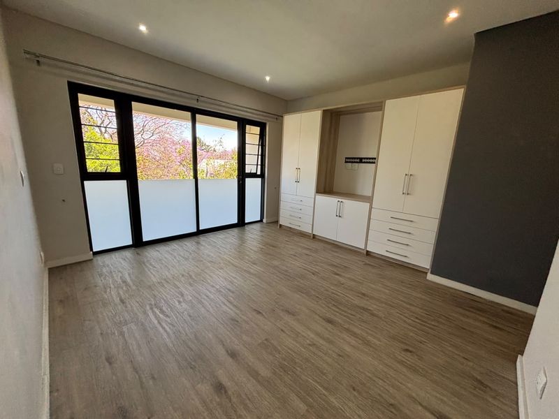 A bright, empty bedroom featuring modern laminate flooring, extensive built-in cabinetry, and large sliding glass doors providing natural light and garden views.