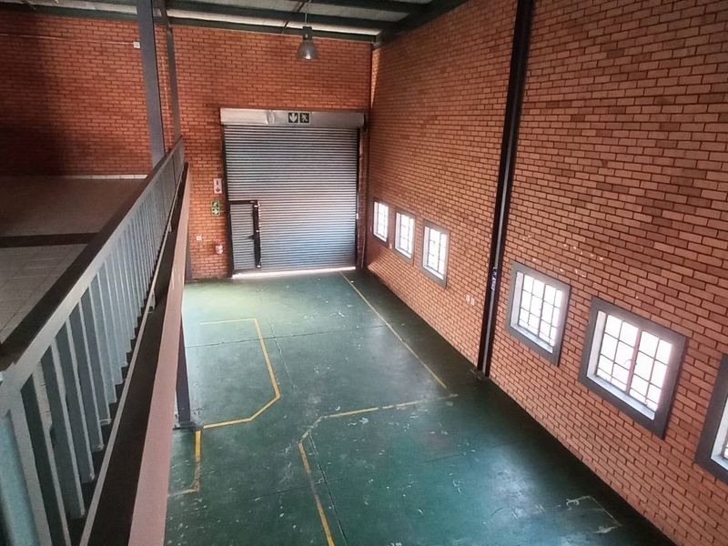 Spacious industrial warehouse unit featuring exposed brick walls, high ceilings, a large roller shutter door, and a mezzanine level with metal railing.